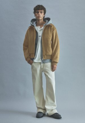 Jeune homme debout portant une veste zippée beige sur un sweat à capuche gris, une chemise blanche, un pantalon beige clair et des chaussures noires sur fond uni.