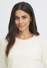 Pull en crème avec un col rond à bords festonnés et de délicats détails en tricot. Texture douce et manches longues visibles.