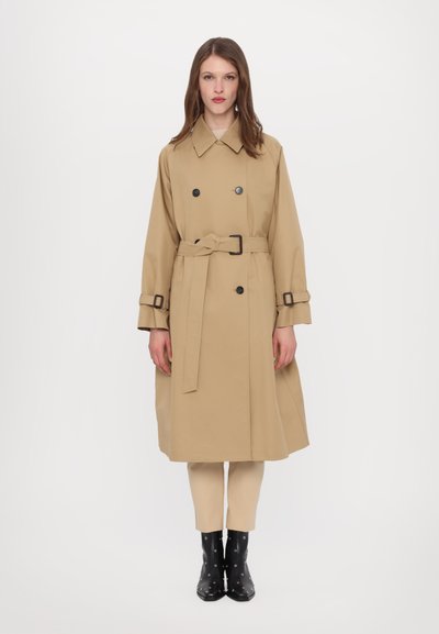 Beige kaksinkertainen trench-coat vyöllä, suuri kaulus, mustat napit ja säädettävät hihansuut; yhdistettynä vaaleisiin housuihin ja mustiin saappaisiin.