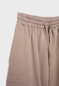 Beige Sweatpants mit elastischem Bund, Kordelzug und Seitentasche, die obere Hälfte der Hose vor einem einfarbigen Hintergrund zeigend.