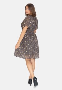 Vestido negro con un patrón floral, hecho de una tela ligera, falda acampanada, mangas cortas abullonadas y detalle de cuello atado. Combinado con tacones negros.