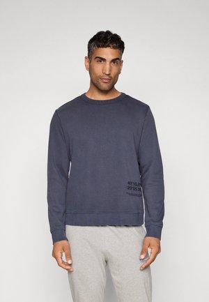 Man with short dark hair wearing a dark blue sweatshirt with printed coordinates and light gray sweatpants, standing against a plain background.
