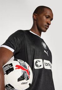 Maillot de football noir avec des accents et un logo blancs ; matériau texturé. Joueur tenant un ballon de football noir et blanc avec une marque rouge.