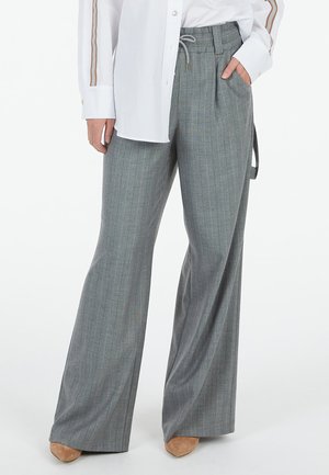 Person wearing wide-leg gray pinstripe trousers with drawstring waist and a white long-sleeve shirt, standing with one hand in pocket.