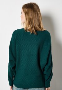 Frau mit schulterlangem blonden Haar, die einen strukturierten dunkelgrünen Pullover und blaue Jeans trägt und einer weißen Wand zugewandt ist.