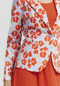 Blazer floral avec une base bleu clair et des fleurs rouges vibrantes. Comporte des revers structurés, une fermeture à un bouton, et un haut côtelé orange en dessous.