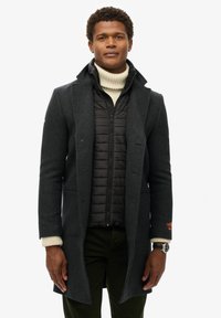 Ciemnoszary wełniany płaszcz na czarnej kurtce puchowej, z wysokim kołnierzem, zapięciem na guziki z przodu oraz widocznymi mankietami o dzianinowej fakturze.