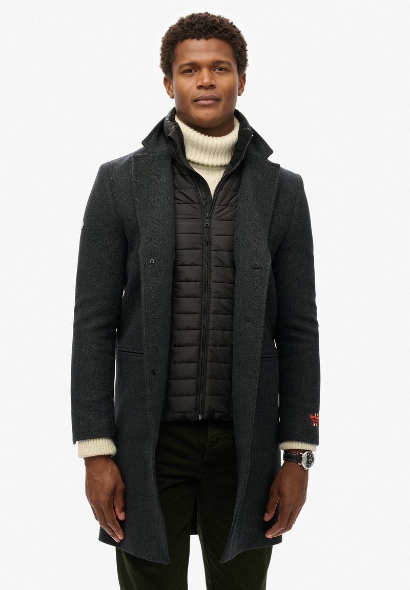 Ciemnoszary wełniany płaszcz na czarnej kurtce puchowej, z wysokim kołnierzem, zapięciem na guziki z przodu oraz widocznymi mankietami o dzianinowej fakturze.