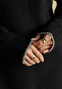 Melns džemperis ar ribotām piedurknēm, kam ir pelēka apdare. Rokas demonstrē zelta gredzenus un kārtīgi koptas nagus, radot teksturētu, kārtotu izskatu.