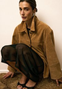 Ruskean mokkanahkainen takki, jossa on kaulus ja napitus, käytetty läpikuultavan mustan mekon päällä. Malli kyykistyy teksturoidulla pinnalla esitellen kenkiään.