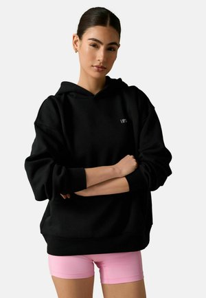 Sort hættetrøje med løs pasform og hætte. Har ribstrikkede ærmegab og forneden samt lille hvid logotekst på fronten. Model iført lyserøde shorts.