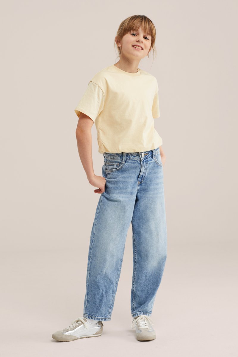 Jeune personne souriante aux cheveux châtain clair portant un T-shirt jaune pâle, un jean large bleu clair et des baskets blanches, debout de manière décontractée.