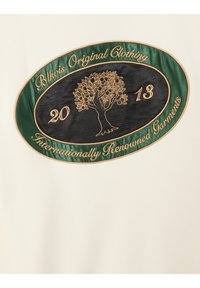 Crèmekleurige stof met een geborduurd logo. Het ontwerp bevat een groene ovale vorm met gouden tekst en een boommotief, met de tekst "Blkvis Original Clothing 2013."