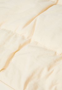 Couverture matelassée beige clair avec une texture douce et lisse, présentant des coutures visibles et un aspect légèrement froissé.
