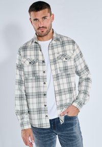 Karierte Flanellhemden in Creme und Schwarz mit zwei Brusttaschen, Knopfverschluss vorne und langen Ärmeln. Über einem weißen T-Shirt getragen.