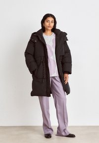 Schwarzer Steppmantel mit hohem Kragen, Reißverschluss und Druckknopfverschluss sowie Fronttaschen. Getragen über einem hellgrauen Sweatshirt und lavendelfarbenen Schlaghosen.