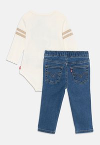 Krämfärgad långärmad babydress med beige ränder på ärmarna, parat med blå denimjeans med elastisk midja, båda visas bakifrån.