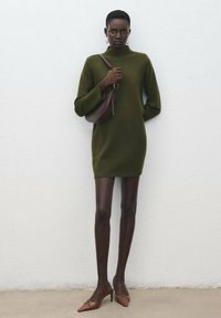 Robe tricotée verte avec un col haut, des manches longues et des détails de boutons dorés. Associée à des escarpins marron à bout pointu et à un petit sac marron.