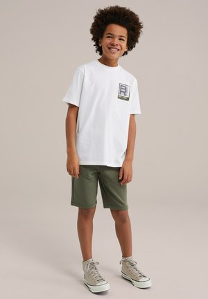 T-shirt bianca in cotone con un design grafico sulla tasca, abbinata a pantaloni corti verde oliva. Il modello indossa sneakers alte grigio chiaro.
