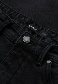 Vaqueros de mezclilla negra con una textura suave. Presenta un botón de metal con "Ombre Denim" grabado y una etiqueta que dice "ombre M32."