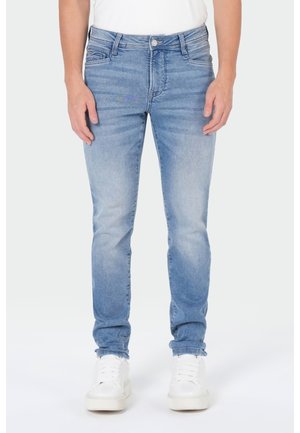 Mann trägt hellblaue Slim-Fit-Jeans und weiße Sneakers, steht mit entspannten Händen neben seinem Körper auf einem einfarbig hellen Hintergrund.