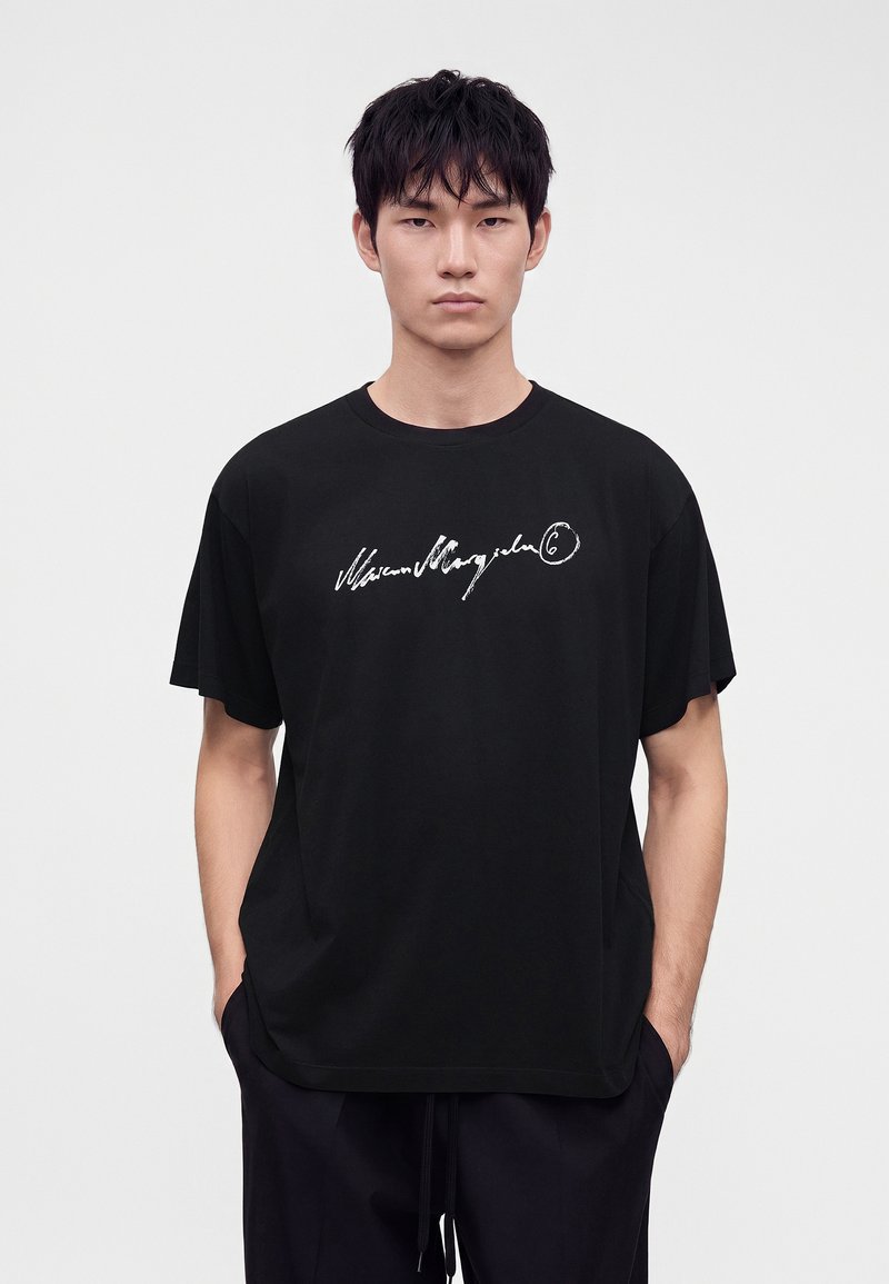 Joven con cabello corto y negro lleva una camiseta negra holgada con texto en cursiva blanca en el frente, de pie contra un fondo gris claro liso.