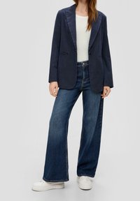 Granatowy blezer z wywiniętym kołnierzem, zestawiony z dżinsami o wysokim stanie i szerokimi nogawkami. Całość dopełniają białe sneakersy na platformie.