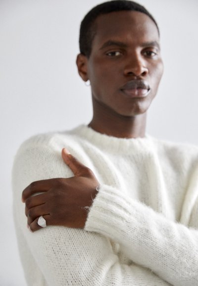White, fluffy knit sweater with ribbed cuffs worn by a person. They are holding their arm, displaying a textured silver ring on their hand.
