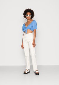 Top cropped azul claro com mangas bufantes e frente com laço, combinado com calças brancas de cintura alta e sandálias brancas chunky com sola preta.