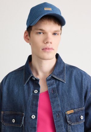 Veste en denim bleu foncé avec des boutons métalliques et une poche latérale, portée par-dessus un débardeur en côte rose, surmontée d'une casquette bleue avec un logo.
