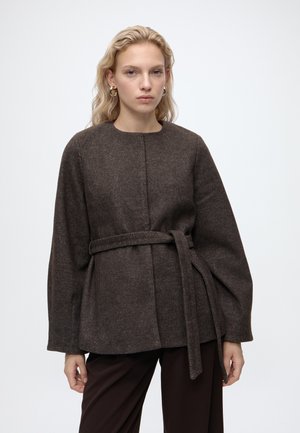 Femme blonde portant un manteau marron foncé ceinturé avec de larges manches et un pantalon foncé, se tenant devant un fond clair uni.