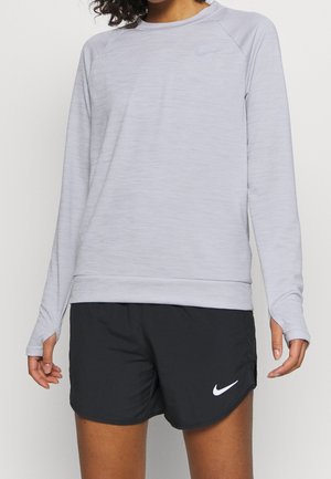 Femme portant un haut de sport à manches longues gris clair avec des trous pour les pouces et un short de course Nike noir avec logo blanc.