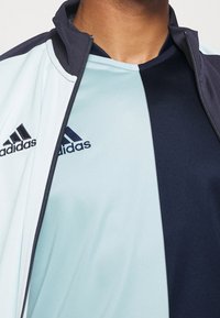 Chaqueta de entrenamiento de Adidas con tejido azul claro, acentos azul oscuro y cierre de cremallera. Logo notable en la parte izquierda del pecho.