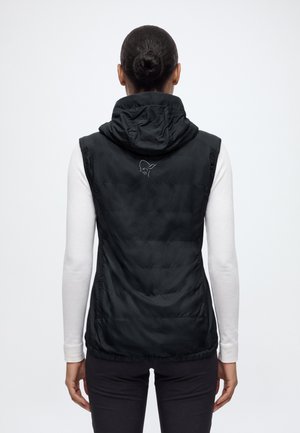Vue de dos d’une personne portant un gilet noir à capuche par-dessus une chemise blanche à manches longues et un pantalon noir, sur un fond uni.