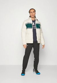 Mand iført en cremefarvet pufferjakke med grøn stribe, navy grafisk T-shirt, sorte træningsbukser og sorte sneakers med blå såler, stående.