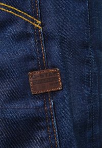 Jeans de mezclilla con tela azul oscura, costuras naranjas y una etiqueta de cuero marrón cosida en la cinturilla que presenta texto en relieve.