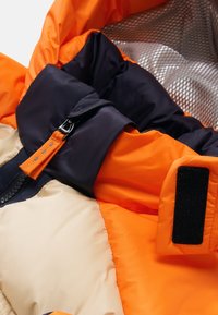 Puffy jacket with orange, cream, and navy segments. Features a zipper, adjustable strap, and textured lining. Velcro closure detail.