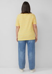 Gelb-weiß gestreifes T-Shirt mit kurzen Ärmeln, getragen zu hellblauen, locker sitzenden Jeans, von hinten gegen einen neutralen Hintergrund gezeigt.