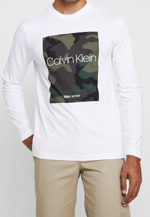 Muž v bílé košili s dlouhým rukávem s maskáčovým čtvercem a nápisem "Calvin Klein 1968 NYNY", v kombinaci s béžovými kalhotami.