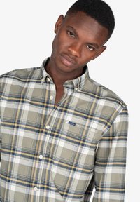 Camisa a cuadros en verde y beige con acentos en azul y amarillo. Cuenta con cuello con botones, mangas largas y un pequeño logotipo de la marca en el pecho.