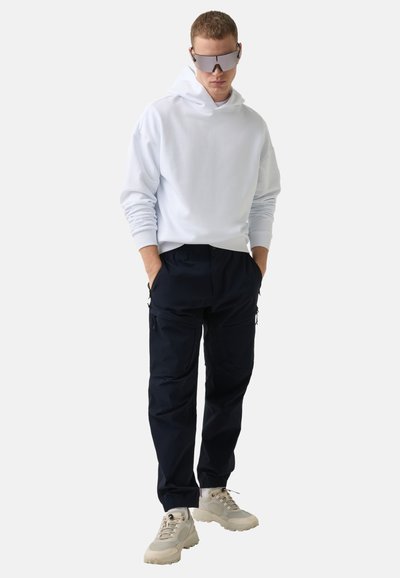 Bílá mikina s kapucí, námořnické cargo kalhoty s kapsami a béžové sneakers. Model nosí reflexní sluneční brýle; hladké textury a uvolněný střih.