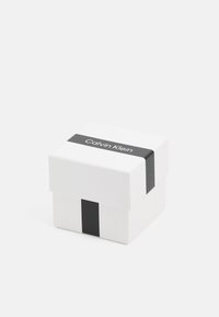 Valkoinen pahvilahjalaatikko, jossa on musta nauha, johon on painettu "Calvin Klein" -logo. Neliskulmainen muoto, sileä pinta, suljettu kansi ja minimalistinen muotoilu.