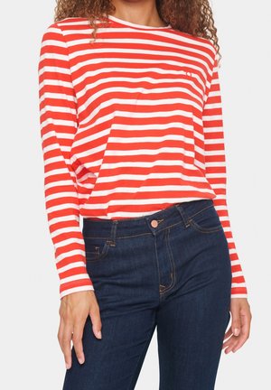 Femme portant un haut à manches longues rayé rouge et blanc rentré dans un jean bleu foncé, sur un fond uni.