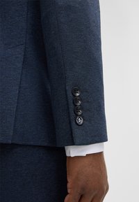 Rękaw granatowej marynarki z fakturowanej tkaniny, z czterema czarnymi guzikami i białym mankietem koszuli wystającym spod spodu.