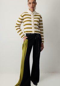 Gestreepte cardigan in crème en olijfgroen met knopen; gecombineerd met zwarte flared broeken en een groene satijnen sjaal. Gebreide textuur zichtbaar.