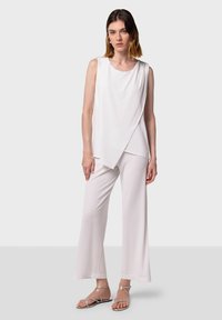 Haut sans manches blanc avec un ourlet asymétrique, associé à un pantalon large et léger. Le mannequin porte des sandales à brides argentées. Tissu lisse et léger.