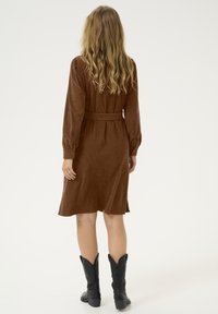 Robe en velours côtelé marron avec des manches longues, une ceinture à nouer à la taille et une jupe fluide. Portée avec des bottes de cowboy noires.