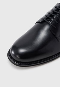 Chaussure habillée en cuir noir poli avec bout arrondi et lacets noirs sur fond blanc.