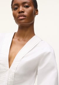 Blouse blanche avec un col en V profond et des accents côtelés texturés le long du col. Présente une coupe ajustée et un tissu en coton doux avec une broderie subtile.