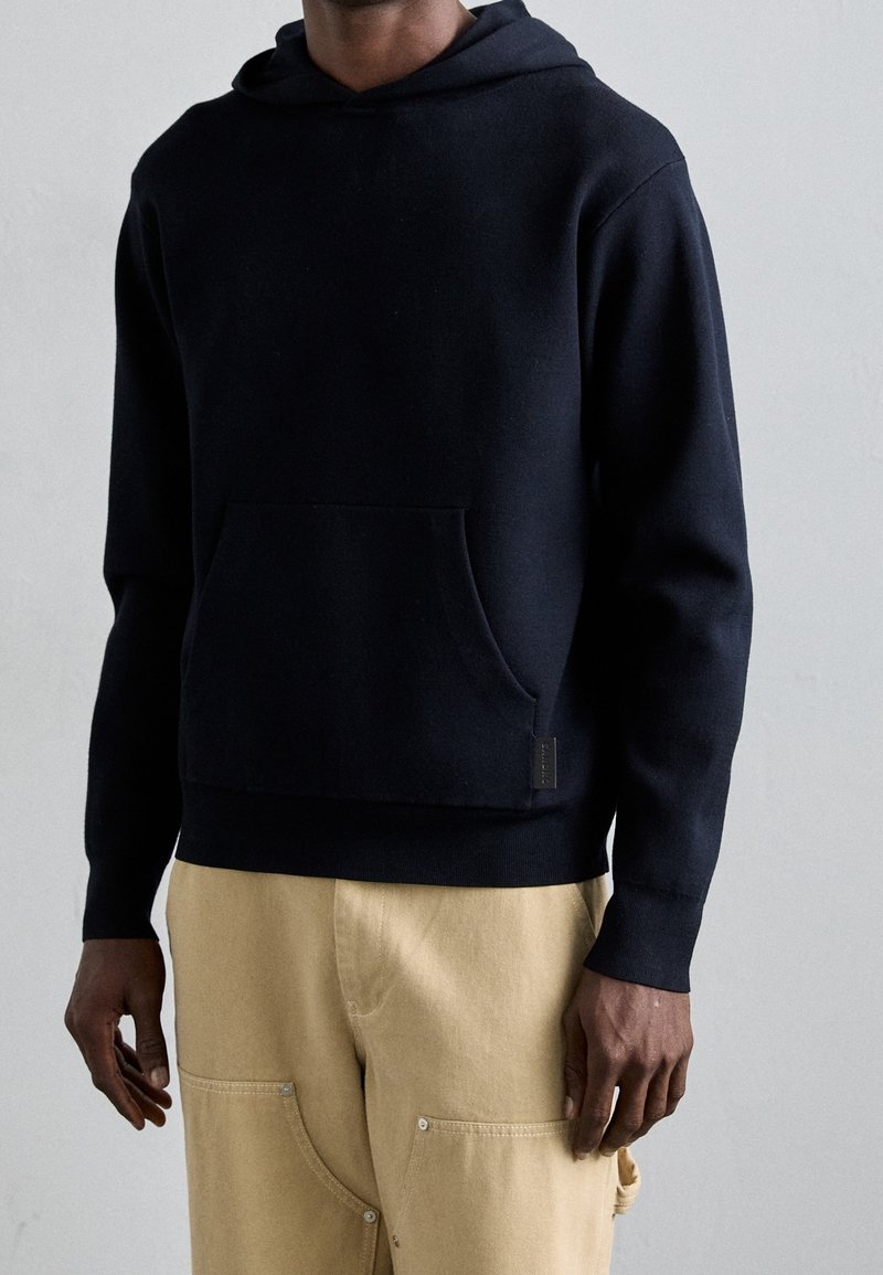 Personne portant un sweat à capuche noir et un pantalon beige, debout devant un fond uni gris clair.
